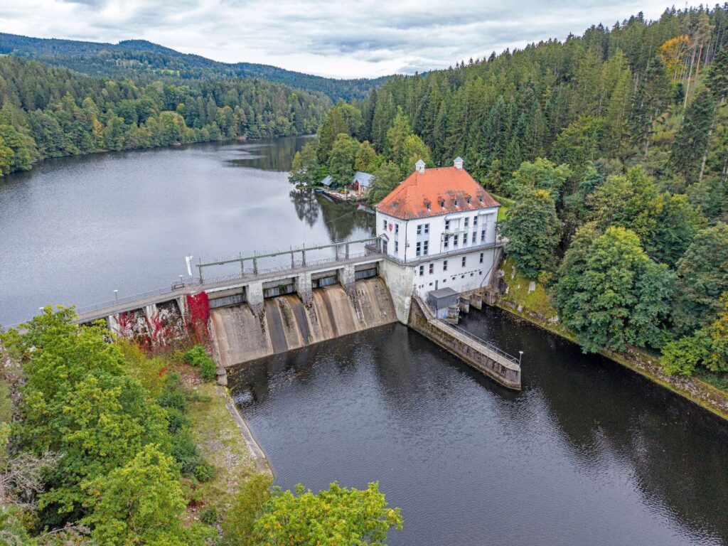 Luftaufnahme vom Wasserkraftwerk Höllenstein mit Staumauer, Betriebsgebäude und Stausee inmitten bewaldeter Hügel im Tal.