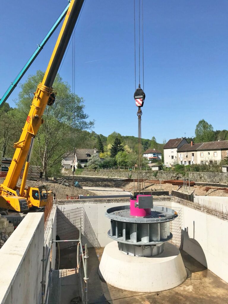 Gelber Mobilkran setzt eine Wasserturbine in eine Betonfassung auf einer Baustelle am Fluss ein.