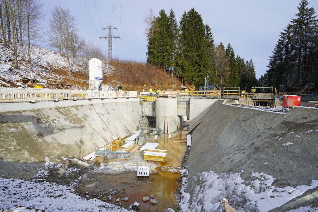 Baustelle für ein Betonbauwerk im Winter mit Silo, Schalung und verschneiten Böschungen am Waldrand.