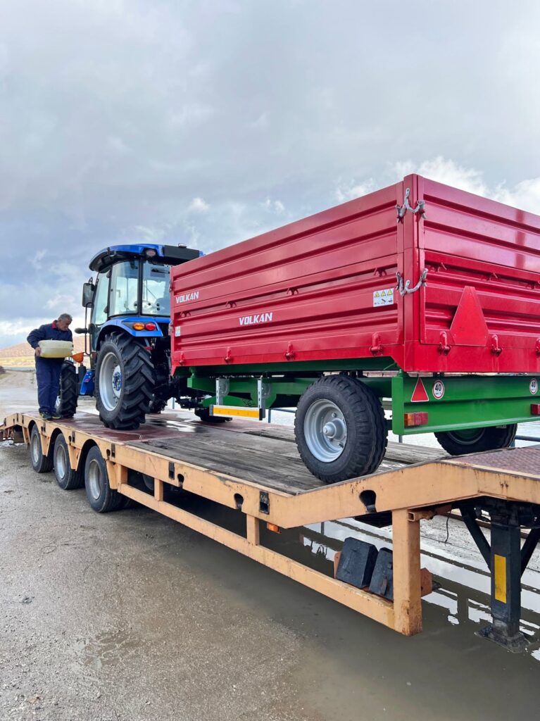 Blauer Traktor mit rotem Volkan-Anhänger auf einem Tieflader-LKW beim Transport im Freien.