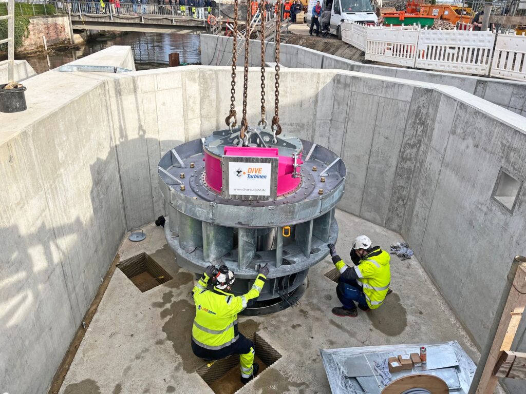 Montage einer DIVE-Turbine in einem Wasserkraftwerk durch zwei Techniker in gelber Warnkleidung auf einer Betonbaustelle.