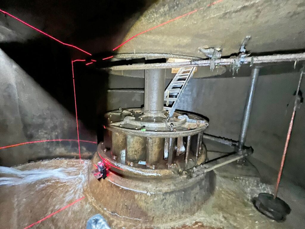 Industrielle Wasserturbine in dunkler Industrieanlage mit roten Laser-Messlinien, Metallleiter und fließendem Wasser.