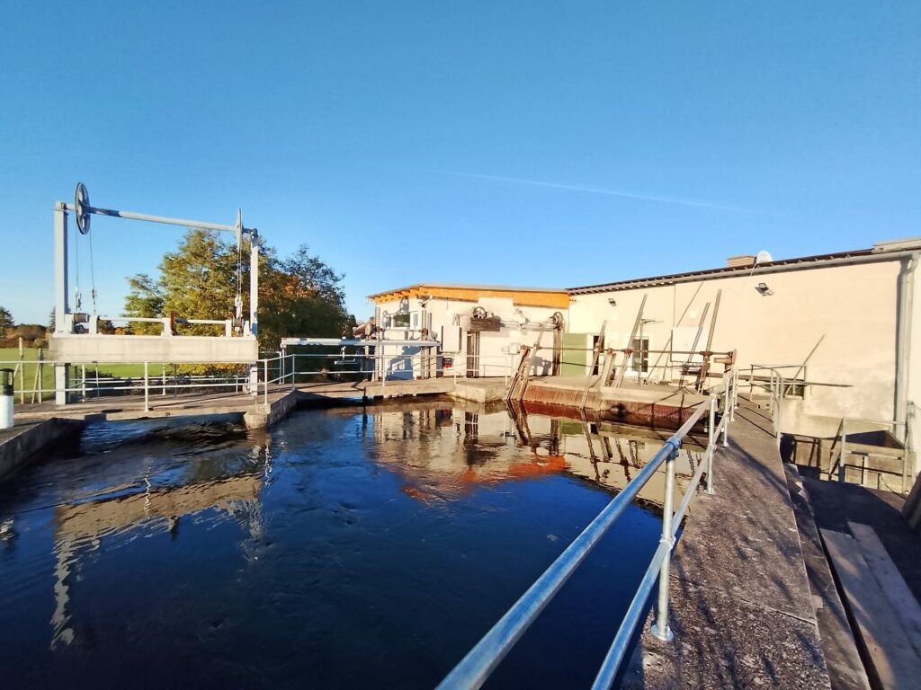 Industrielle Wehranlage an einem Flusslauf mit Rechenreiniger und Betriebsgebäude unter blauem Himmel bei Sonnenschein.