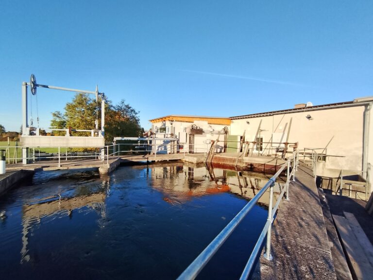 Industrielle Wehranlage an einem Flusslauf mit Rechenreiniger und Betriebsgebäude unter blauem Himmel bei Sonnenschein.