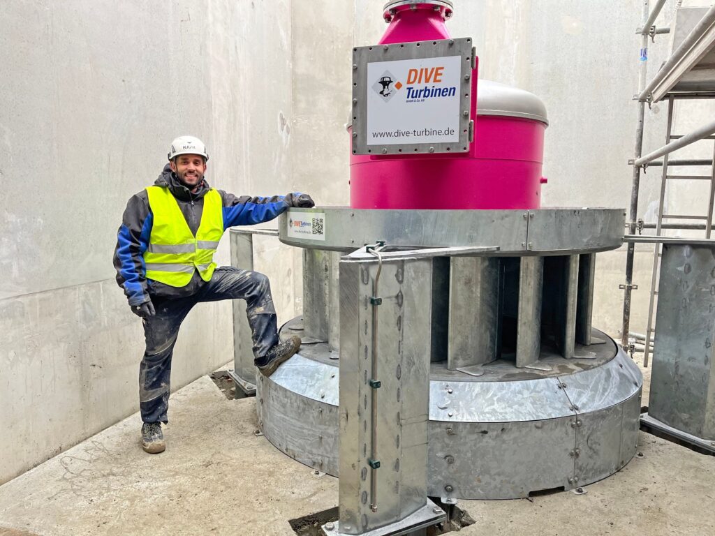 Monteur in Warnweste neben einer großen, pinkfarbenen DIVE-Turbine in einem Betonschacht auf einer Baustelle.
