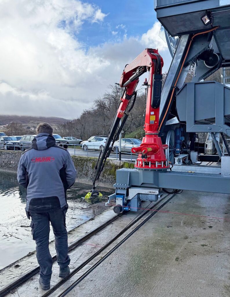 Arbeiter von Muhr bedient einen roten Ladekran am Hafenbecken zur Reinigung des Wasserlaufs an einer Industrieanlage.