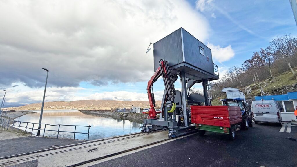Stationäre Rechenreinigungsanlage mit rotem Kranarm und Volkan-Anhänger an einem Wasserlauf vor Hügellandschaft.
