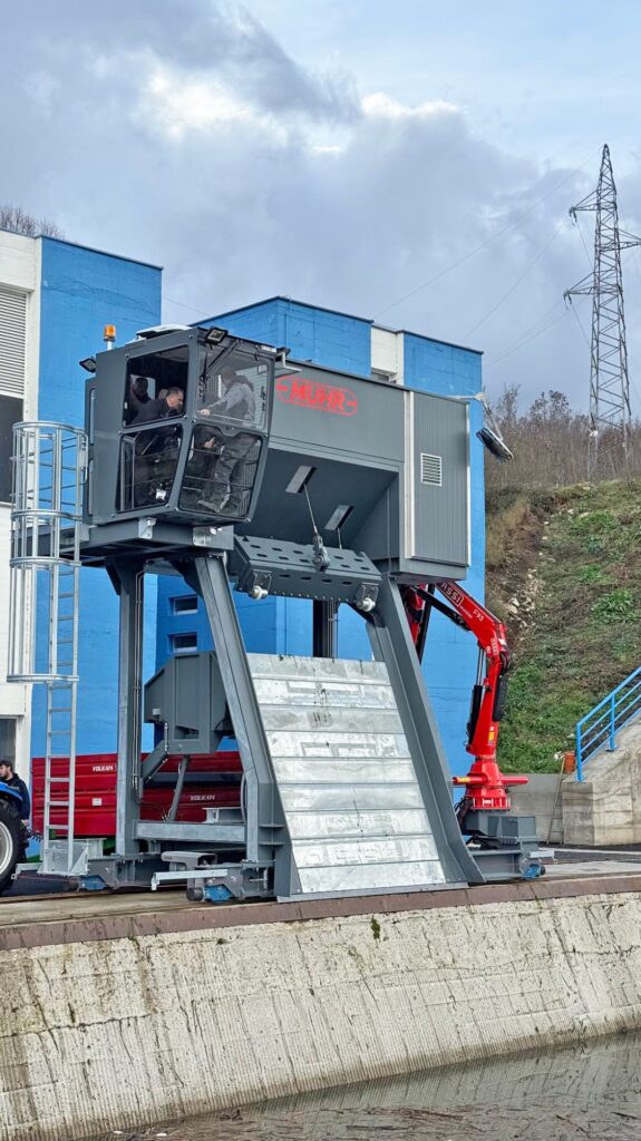 Stationäre Muhr Rechenreinigungsmaschine mit Steuerkabine über einem Wasserkanal vor blauem Industriegebäude.