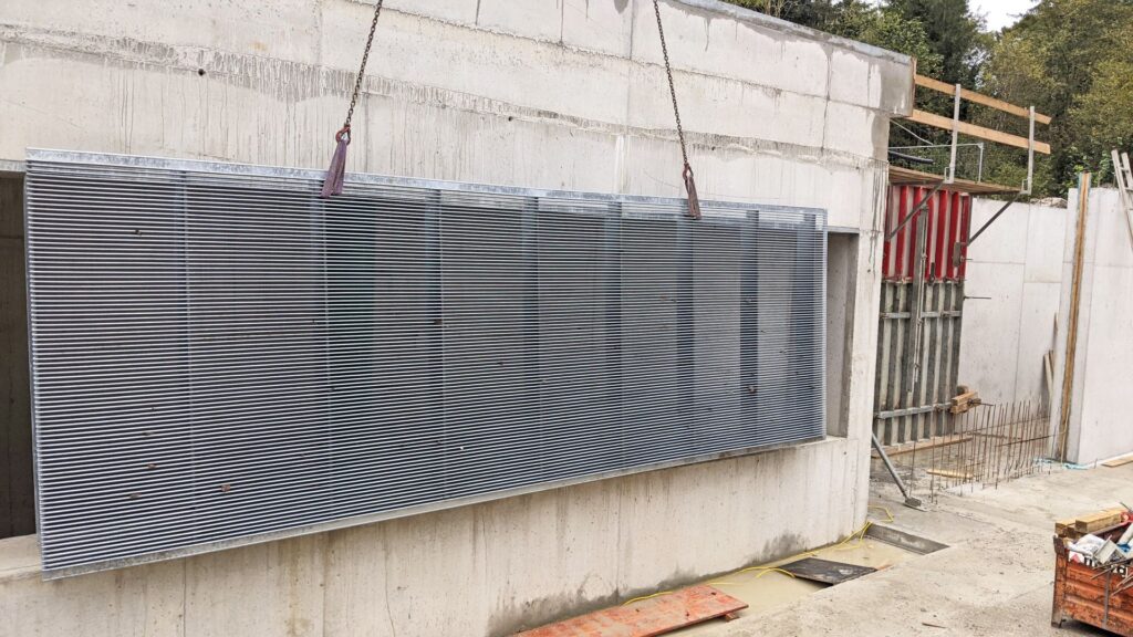 Ein Kran hebt ein großes Gitterrost-Element vor eine Betonwand auf einer Baustelle.