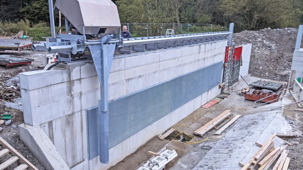 Horizontale Rechenreinigungsanlage an einer Betonmauer auf einer Baustelle für Wasserbau und Infrastruktur.