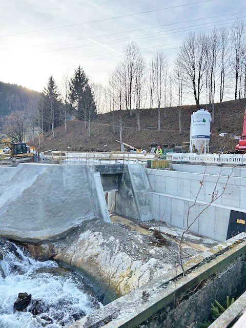 Baustelle am Vilsfall mit Betonmauern, Wasserlauf, Bagger und Silo vor bewaldetem Hang.