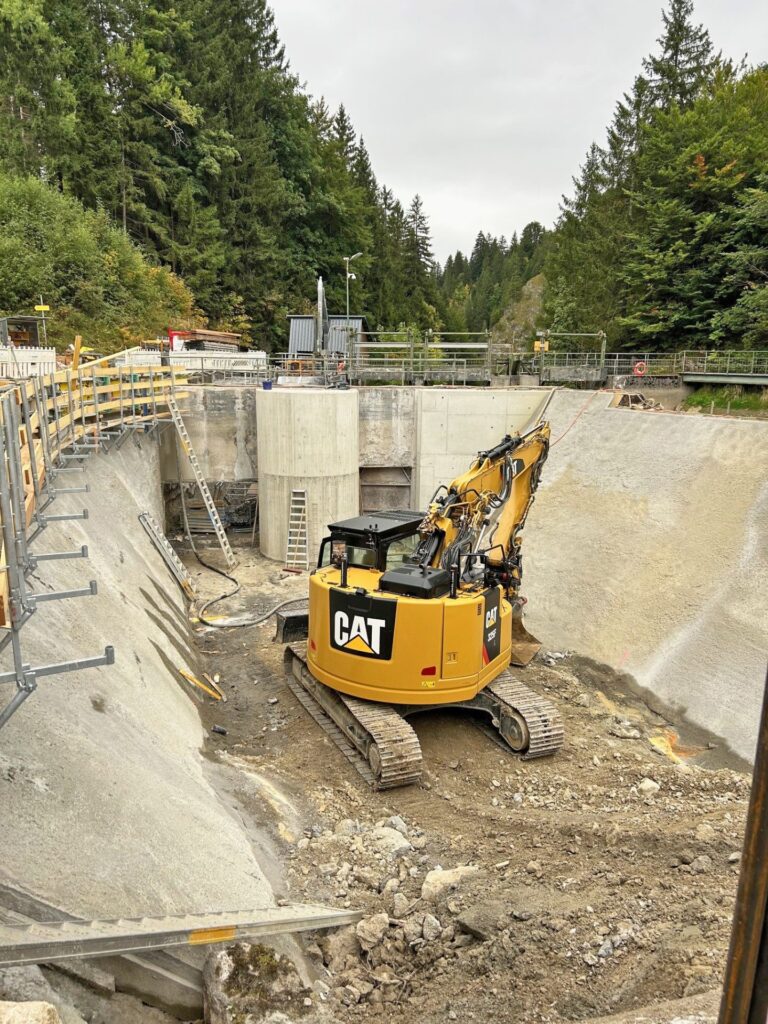 Gelber CAT Kettenbagger bei Bauarbeiten am Vilsfall in einer tiefen Beton-Baugrube vor bewaldeter Kulisse.