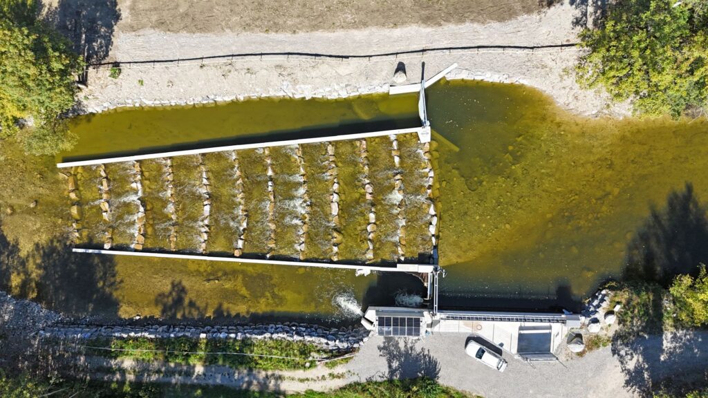 Luftaufnahme einer Wehranlage mit integrierter Fischtreppe aus Steinriegeln und Solarmodulen am Flussufer.