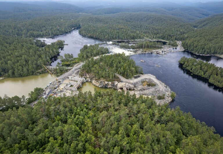 Luftaufnahme einer Wasserkraftwerk-Baustelle am Fluss Fin, umgeben von dichtem Nadelwald und felsiger Landschaft.