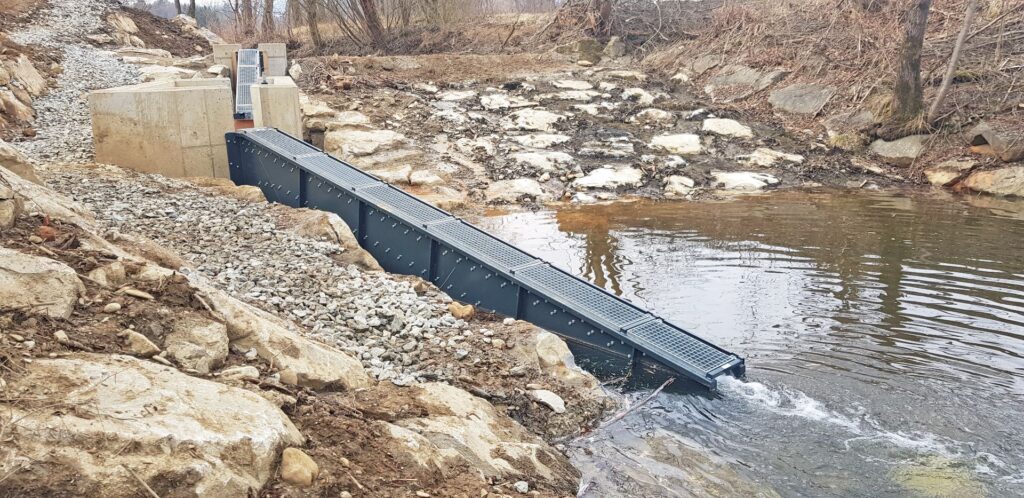 Graue Fischtreppe aus Metall mit Gitterrost-Abdeckung führt von Betonwänden in ein natürliches Fließgewässer.