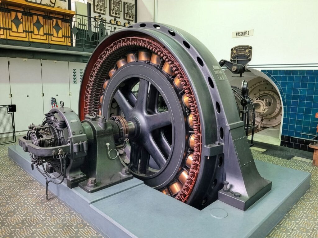 Historischer elektrischer Generator mit großem Schwungrad und Kupferwicklungen in einer Industriehalle.