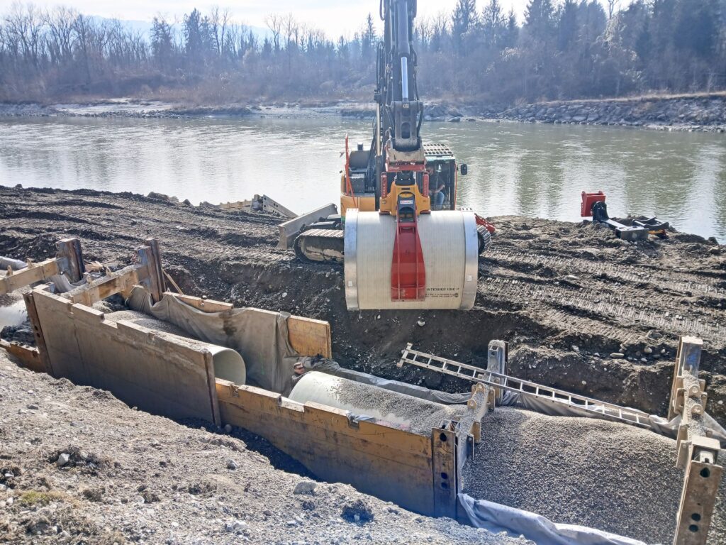 Bagger setzt großes Betonrohr in einen Graben am Flussufer ein, Tiefbauarbeiten mit Verbau und Kiesschüttung.