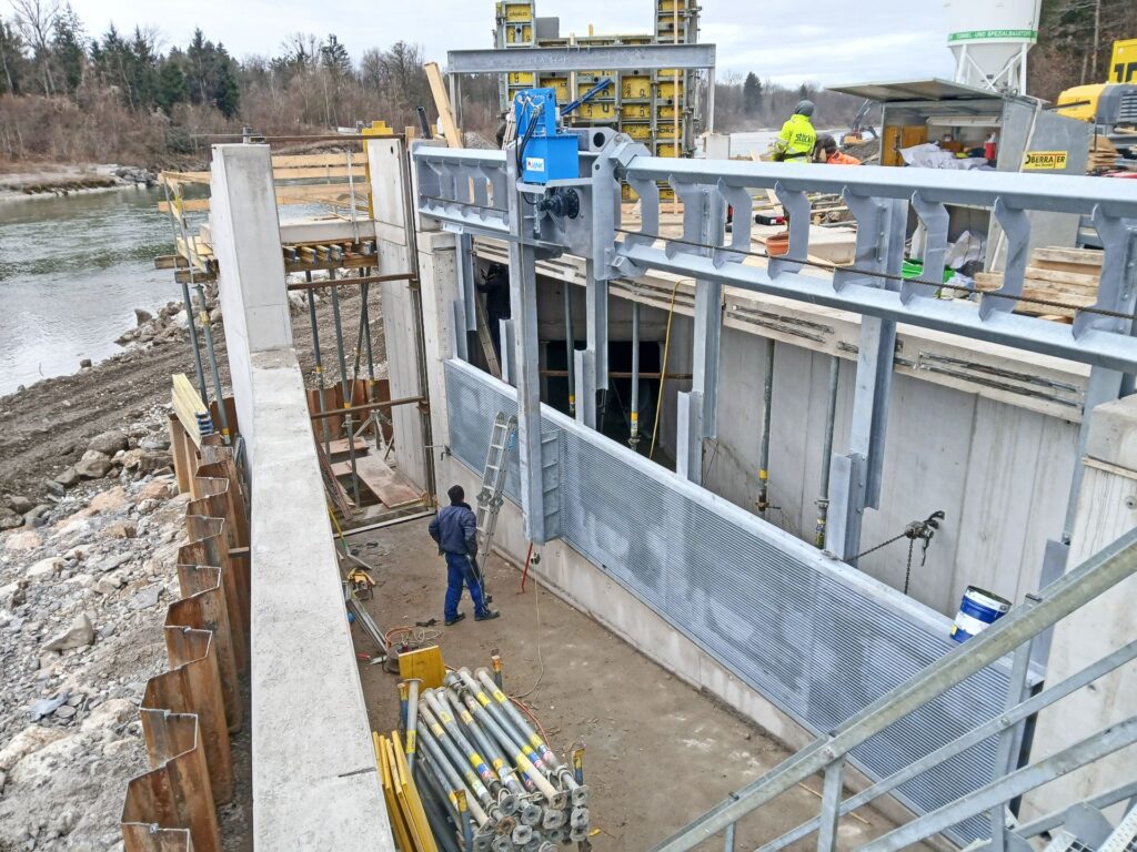 Bauarbeiter an einer Wasserbau-Baustelle mit Spundwänden, Stahlkonstruktion und Rechenreiniger am Flussufer.