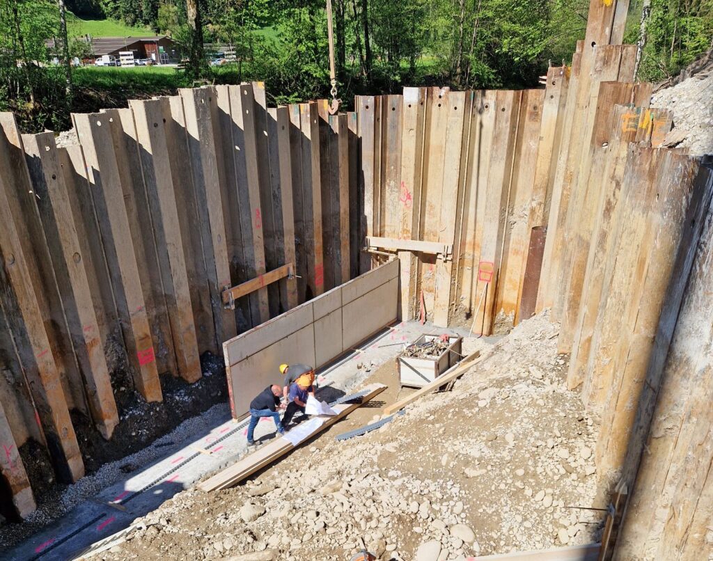 Bauarbeiten in tiefer Baugrube mit Spundwänden aus Stahl, Arbeiter prüfen Baupläne auf dem sandigen Fundament.