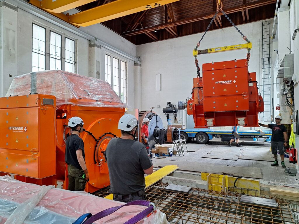 Arbeitertrupp montiert orangefarbenen Hitzinger Generator in Industriehalle; Krantransport von Schwerlast-Equipment.