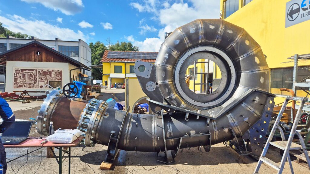 Industrielles spiralförmiges Turbinengehäuse aus Stahl auf einem Werksgelände vor einem gelben Firmengebäude.