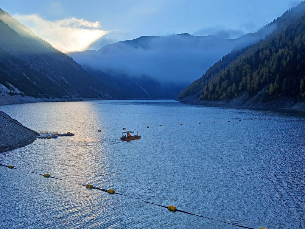 Oranges Boot auf blauem Bergsee im Morgengrauen, umgeben von bewaldeten Hängen und Nebel in den Alpen.
