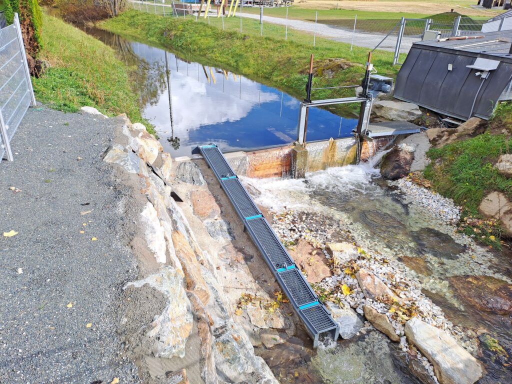 Kleines Wasserkraftwerk mit Rechenreiniger und Fischtreppe an einem Bachlauf mit herbstlicher Uferböschung.