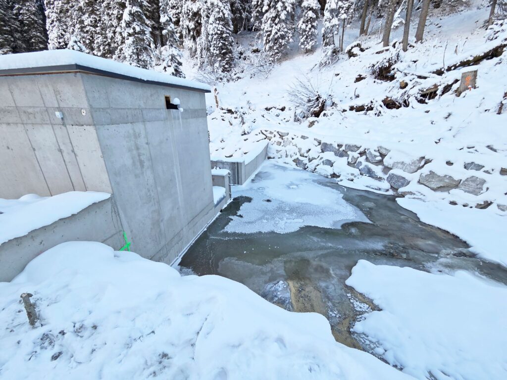 Betongebäude an einem zugefrorenen Bachlauf im verschneiten Wald, Winterlandschaft mit Tannen und Steinmauer.