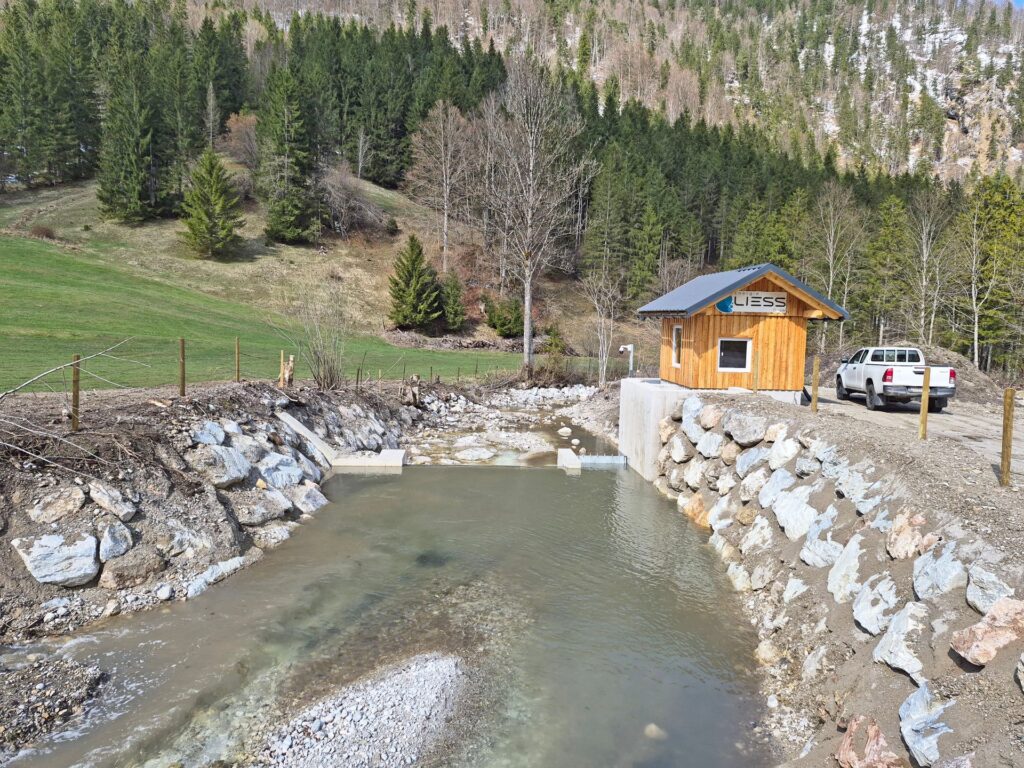 Kleinkraftwerk von Energie LIESS an einem Gebirgsbach mit Holzhaus und Wasserfassung vor bewaldetem Hang.