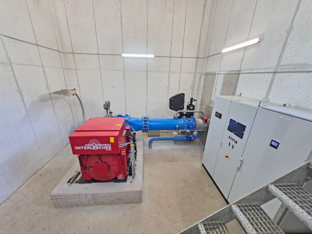 Rote Wasserkraft-Turbine von Maschinenbau Unterlercher mit blauem Rohrleitungssystem und Schaltschrank im Betonraum.