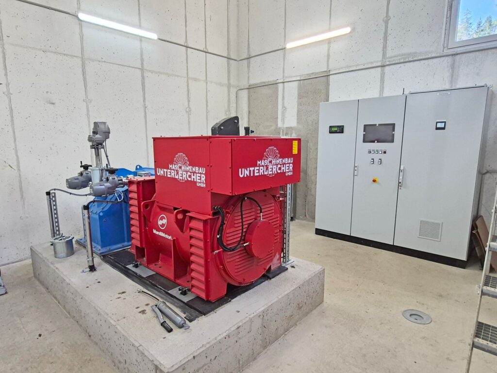 Roter Maschinenbau Unterlercher Generator neben einem grauen Schaltschrank in einer Industriehalle aus Sichtbeton.