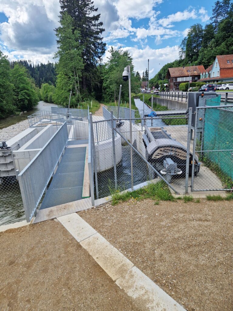 Moderne Fischtreppe aus Beton an einem Flusslauf mit Metallgitterzäunen, Überwachungskamera und dörflicher Kulisse.