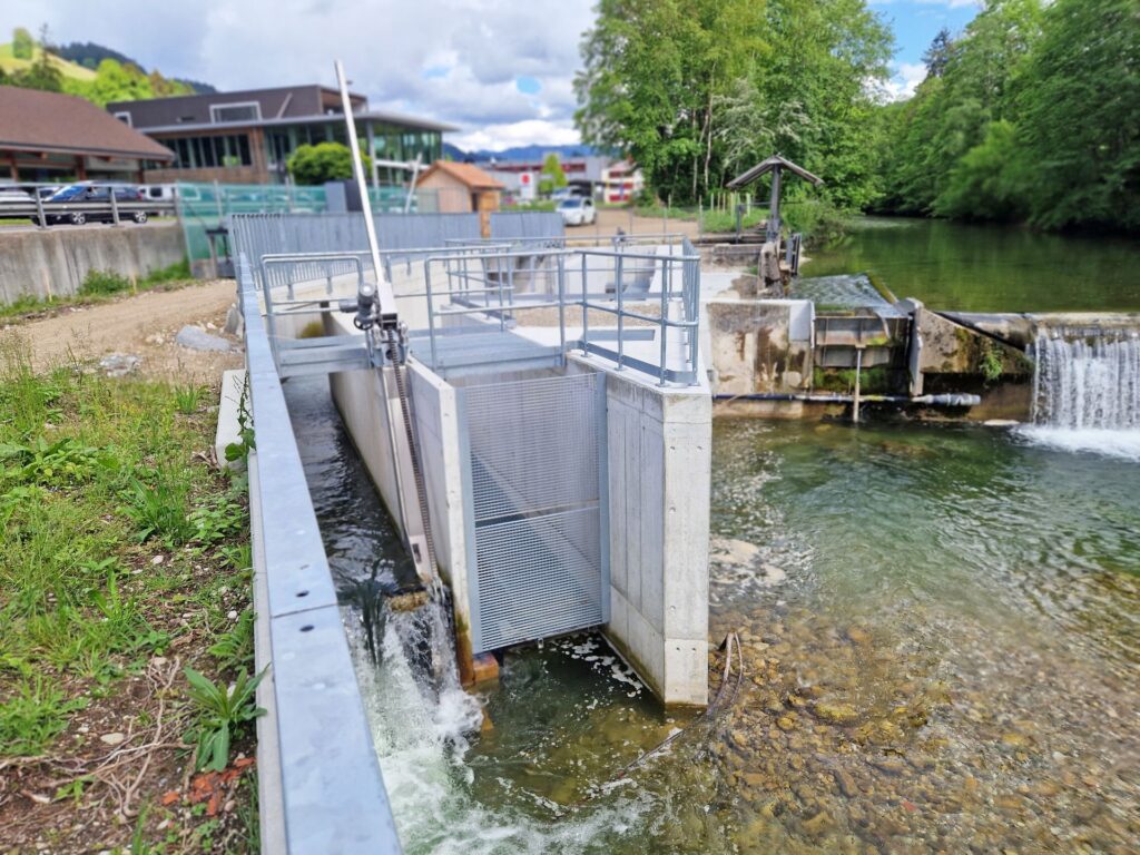 Modernes Beton-Wehr mit Fischtreppe und Rechenanlage an einem Flusslauf vor bewaldeter Hügelkulisse.