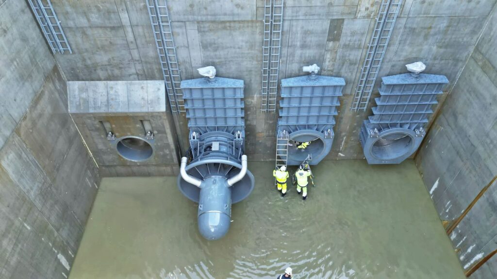 Installierte Wasserturbine und Schütze in einem Betonbecken mit Arbeitern in Warnkleidung zur Wartung.