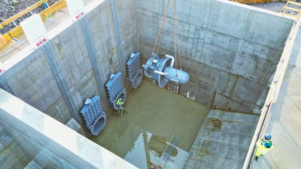 Einbau einer Voith Wasserkraft-Turbine per Kran in ein Betonbecken mit Metallschleusen auf einer Großbaustelle.