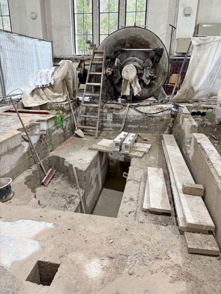 Industrie-Baustelle in einer Halle mit großer Metallturbine, Fundament-Ausgrabungen, Leiter und Werkzeugen auf Betonboden.
