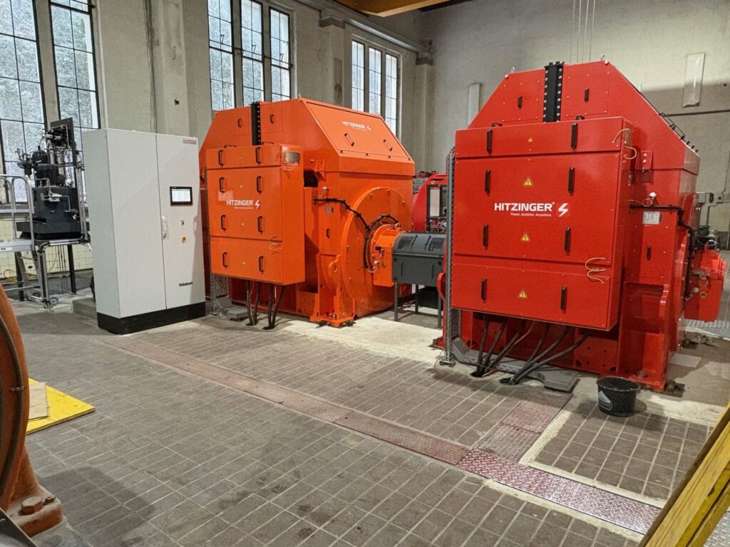 Zwei große, orangefarbene Hitzinger Generatoren in einer Industriehalle neben einem grauen Schaltschrank.