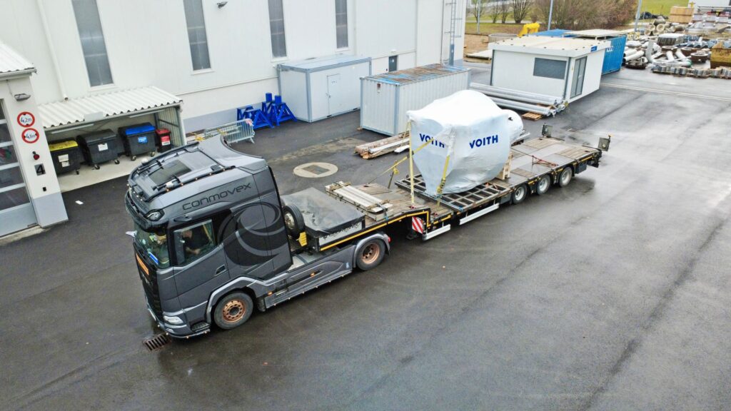 Schwertransport von Conmovex mit verpackter Voith-Maschine auf einem Tieflader vor einem Industriegebäude.