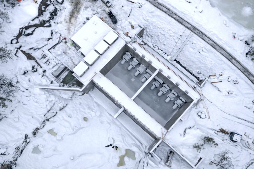 Vogelperspektive auf eine Wasserkraftwerk-Baustelle im Schnee mit Turbinenbecken und schneebedeckten Fundamenten.
