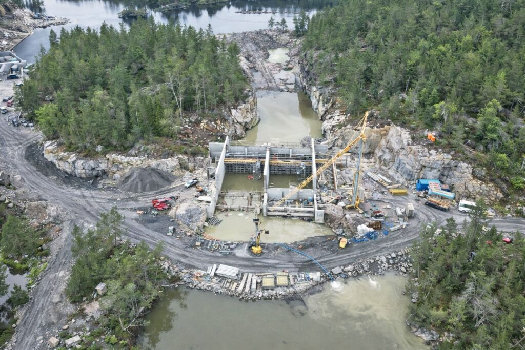 Luftaufnahme der Baustelle einer Staumauer mit gelbem Kran inmitten einer bewaldeten Felslandschaft am Flusslauf.