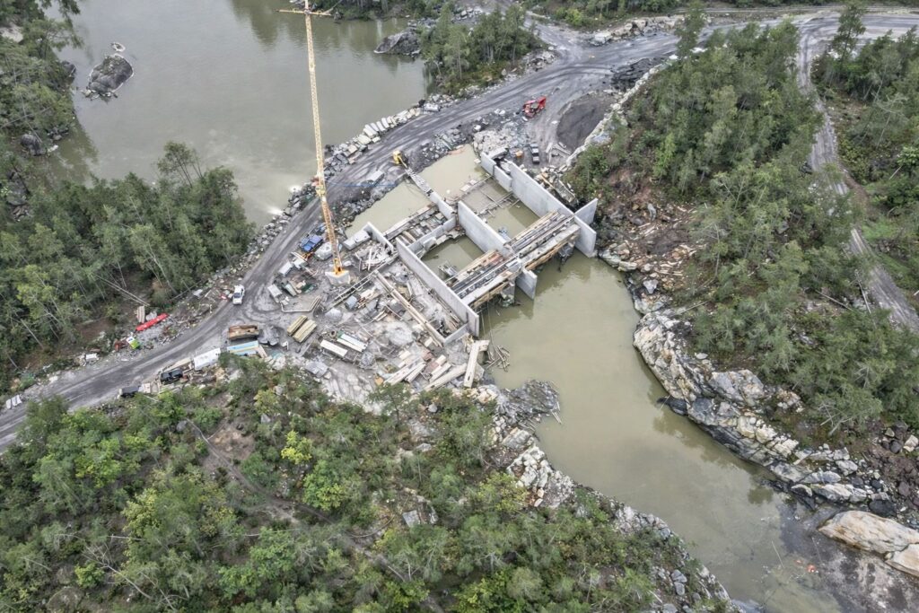 Luftaufnahme einer Baustelle für ein Wehr oder Kraftwerk an einem bewaldeten Flussufer mit Baufahrzeugen und Kran.