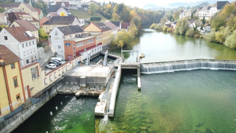 Luftaufnahme eines Flusswehres mit Fischtreppe, angrenzenden bunten Stadthäusern und herbstlichen Bäumen am Ufer.