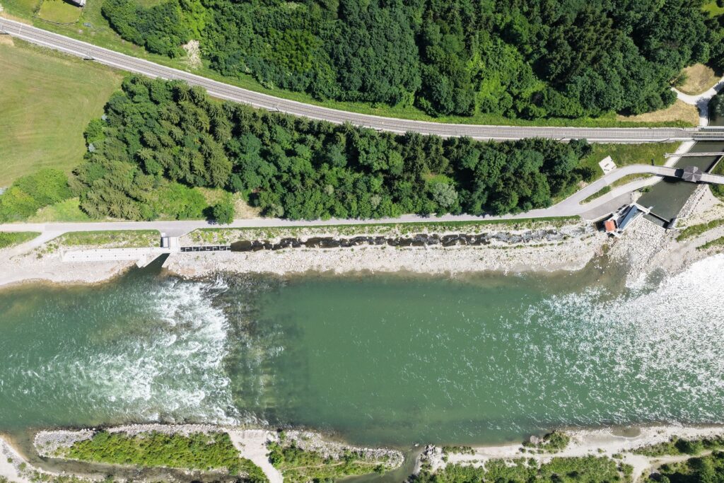 Luftaufnahme eines Wehrs im Fluss mit Fischtreppe, angrenzenden Bahngleisen und dichtem Wald am Ufer.