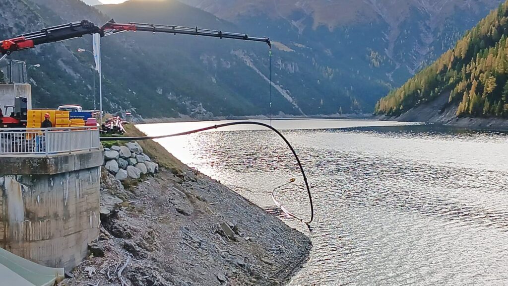 LKW-Kran installiert eine schwarze Förderleitung an einem Gebirgsstausee zwischen steilen Berghängen.