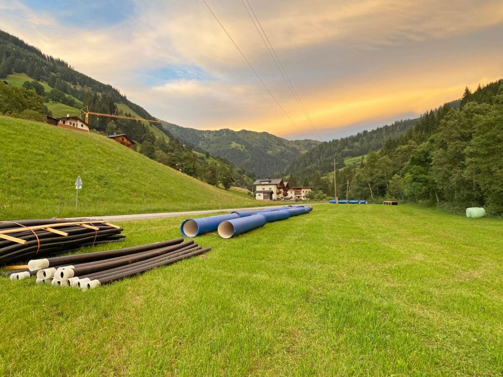 Blaue und schwarze Rohre auf einer grünen Wiese in den Alpen bei Sonnenuntergang vor Berghängen und Häusern.