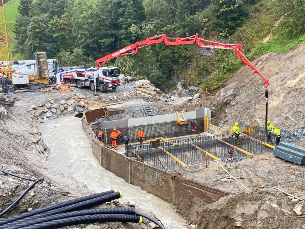 Bauarbeiter betonieren Brückenfundament am Fluss mit roter Betonpumpe und LKW an einer Waldbaustelle im Gebirge.
