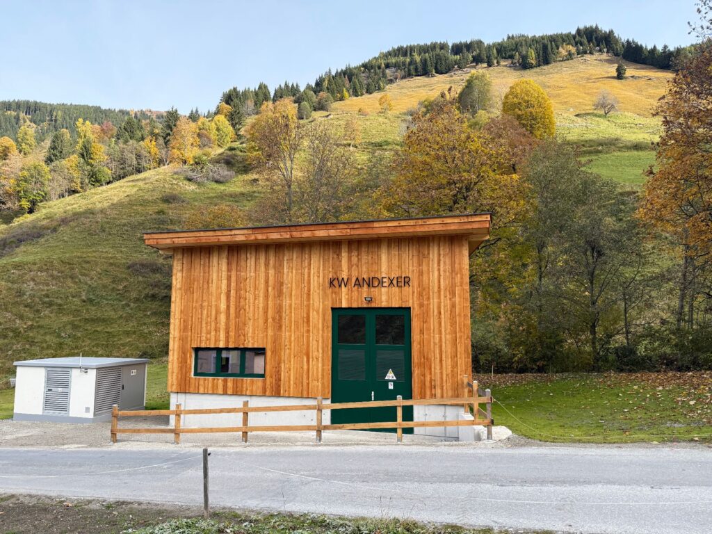 Kraftwerk Andexer aus Holz mit Pultdach vor einem bewaldeten Berghang in herbstlicher Landschaft.