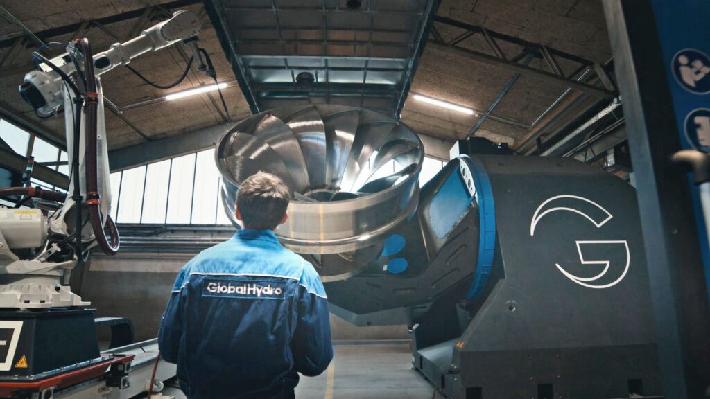 Automatisierte Schweißanlage bei Global Hydro mit Roboterarm und Techniker vor einer großen Wasserturbine in der Fertigung.