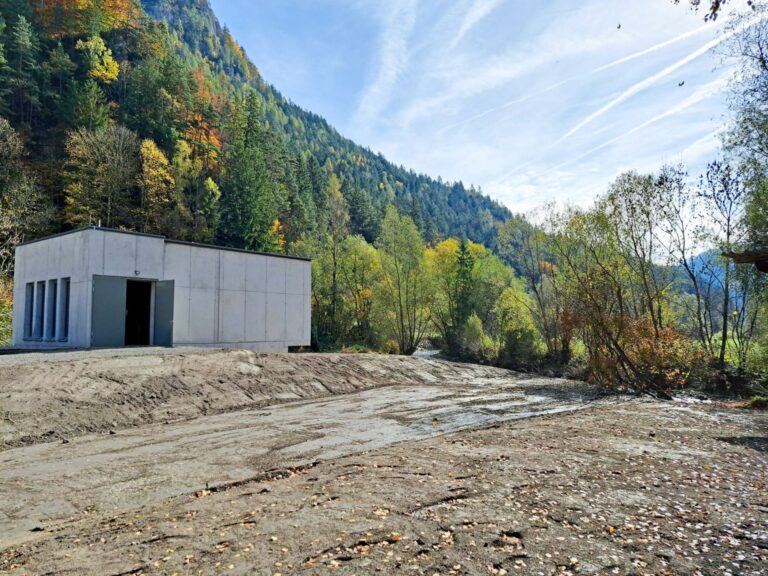 Moderner Betonbau der Hansenhütte am bewaldeten Berghang mit herbstlichen Bäumen und geschotterter Fläche davor.