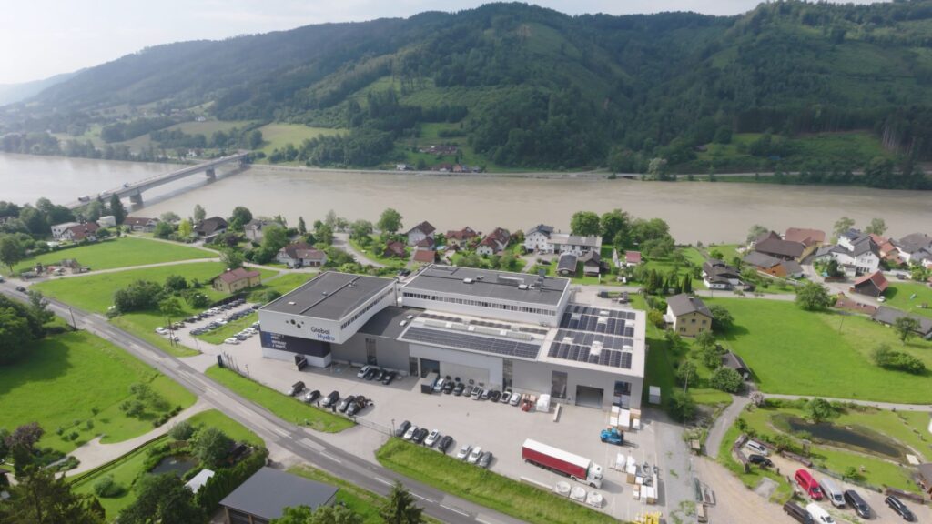 Luftaufnahme des Global Hydro Headquarters in Österreich mit Solaranlagen am Flussufer der Donau vor bewaldeten Hügeln.